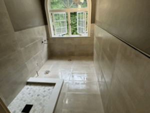 Bathroom Tile Installation in Progress – Wide view of the shower area, showcasing beige tiles, a large window, and unfinished floor installation.