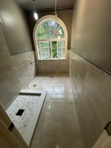 Bathroom Tile Installation in Progress – Wide view of the shower area, showcasing beige tiles, a large window, and unfinished floor installation.